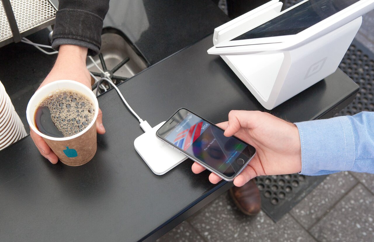 A traveler taps an iPhone showing Apple Pay against a Square contactless reader at a café